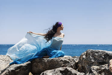 Tänzerin mit Schal auf einem Felsen am Meer sitzend an einem sonnigen Tag - MRRF00881