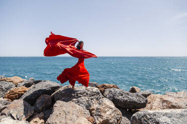 Reife Tänzerin in rotem Kleid tanzt mit Sarong auf Felsen am Meeresufer - MRRF00883