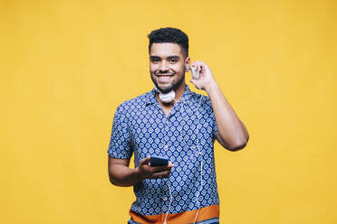 Lächelnder gut aussehender Mann, der Kopfhörer trägt und ein Handy vor gelbem Hintergrund hält - DGOF01948