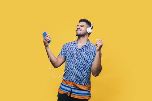 Cheerful man with mobile phone dancing while listening music through headphones against yellow background - DGOF01951