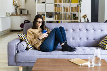 Junge Frau, die ein Mobiltelefon benutzt, während sie sich zu Hause auf dem Sofa ausruht - GIOF11171
