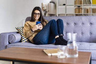 Smiling woman using mobile phone while relaxing on sofa at home - GIOF11172