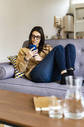 Woman wearing eyeglasses using mobile phone while sitting on sofa at home - GIOF11173