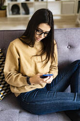 Junge Frau mit Brille, die ein Mobiltelefon benutzt, während sie zu Hause auf dem Sofa sitzt - GIOF11174