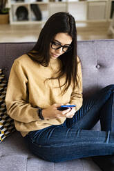 Young woman wearing eyeglasses using mobile phone while sitting on sofa at home - GIOF11174