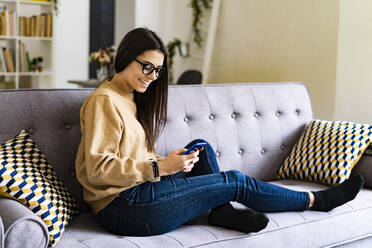 Lächelnde junge Frau, die ein Smartphone benutzt, während sie zu Hause auf dem Sofa sitzt - GIOF11175