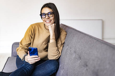 Smiling woman with hand on chin using smart phone while sitting on sofa at home stock photo