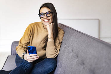 Lächelnde Frau mit Hand am Kinn, die ein Smartphone benutzt, während sie zu Hause auf dem Sofa sitzt - GIOF11176