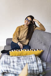 Woman wearing eyeglasses smiling while sitting on sofa at home - GIOF11177