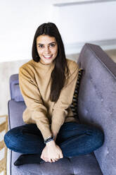 Young woman smiling while sitting cross-legged on sofa at home - GIOF11179
