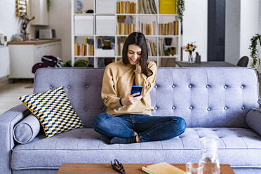 Junge lächelnde Frau, die ein Mobiltelefon benutzt, während sie zu Hause auf dem Sofa sitzt - GIOF11180