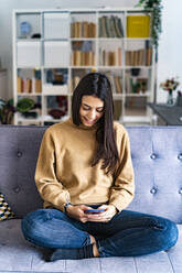 Frau, die ein Mobiltelefon benutzt, während sie im Schneidersitz auf dem Sofa zu Hause sitzt - GIOF11181