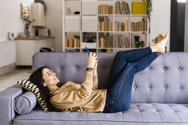 Young woman using mobile phone while lying on sofa at home - GIOF11186