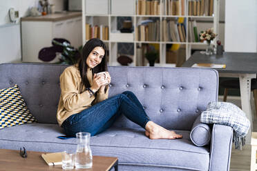 Smiling woman drinking coffee while sitting on sofa at home - GIOF11187