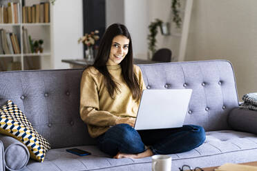 Young woman smiling while using laptop sitting at home - GIOF11191