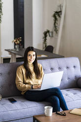 Smiling woman using laptop while sitting with legs crossed at knee on sofa at home - GIOF11192