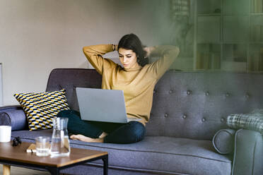 Junge Frau mit Hand im Haar, die auf einen Laptop schaut, während sie zu Hause sitzt - GIOF11195