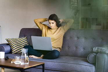 Young woman with hand in hair looking at laptop while sitting at home - GIOF11195