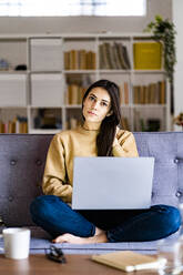 Young woman staring while sitting wit laptop at home - GIOF11196