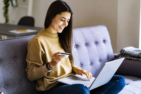 Young woman with credit card shopping over laptop while sitting at home - GIOF11197