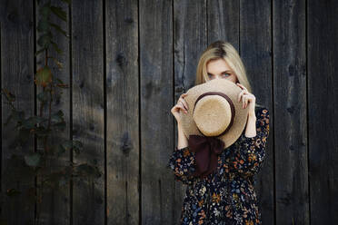 Blonde Frau mit blauen Augen hält sich einen Strohhut vor das Gesicht an einer alten Holzwand - AZF00188