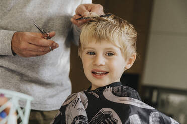 Blond boy smiling when getting haircut from father at home - MFF07133