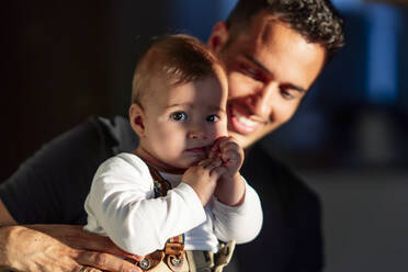 Smiling father holding cute baby boy while standing at home - PGF00437