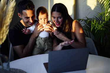 Lächelndes Paar, das seinen Sohn im Arm hält, während es sich über einen Laptop zu Hause per Videoanruf unterhält - PGF00449