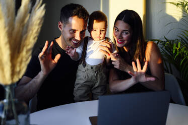 Paar winkt mit der Hand zum Videoanruf auf dem Laptop, während es mit dem Baby zu Hause sitzt - PGF00450