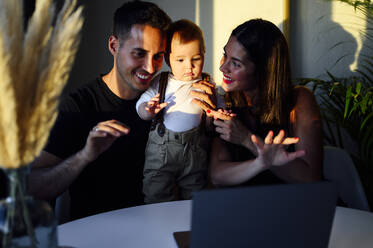 Parents with baby boy waving hand to video call on laptop while sitting at home - PGF00451