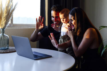 Smiling parents with son waving hand to video call while sitting at home - PGF00452