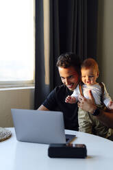 Mid adult man holding baby boy while working on laptop at home - PGF00453