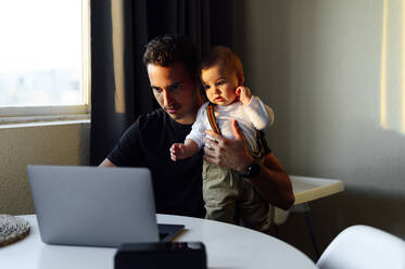 Father holding baby boy while working on laptop at home - PGF00454
