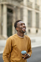 Lächelnder junger Mann mit Einwegbecher, der wegschaut, während er in der Stadt steht - OCMF02064
