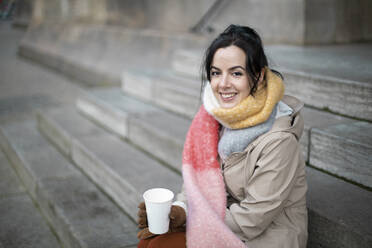 Lächelnde Frau, die im Winter auf einer Treppe sitzend Kaffee trinkt - AXHF00171