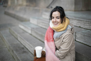 Smiling woman having coffee while sitting on steps during winter - AXHF00171