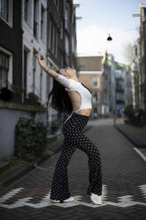 Woman dancing with arms outstretched on footpath at street - AXHF00177