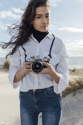 Schöne junge Frau hält Kamera, während sie am Strand gegen den Himmel steht - JRVF00253