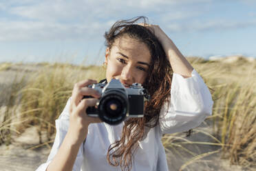 Junge Frau mit Hand im Haar, die eine Kamera hält, während sie vor einem bewölkten Himmel sitzt - JRVF00258