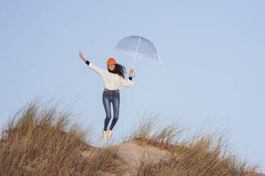 Junge Frau mit Regenschirm springt auf eine Sanddüne - JRVF00264