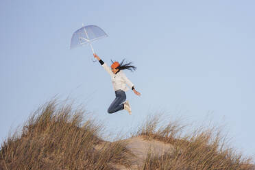 Junge Frau mit Strickmütze springt mit Regenschirm auf einer Sanddüne gegen den Himmel - JRVF00265