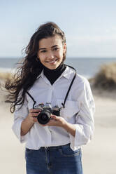 Lächelnde schöne Frau mit Vintage-Kamera steht am Strand gegen den Himmel - JRVF00272