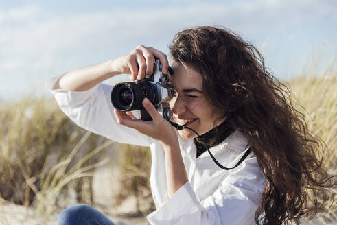 Junge Frau mit langen Haaren beim Fotografieren durch eine alte Kamera am Strand an einem sonnigen Tag - JRVF00273