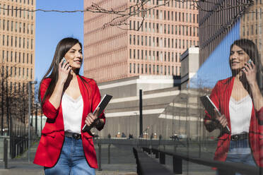 Smiling businesswoman with laptop talking on smart phone while looking at reflection - PNAF00627