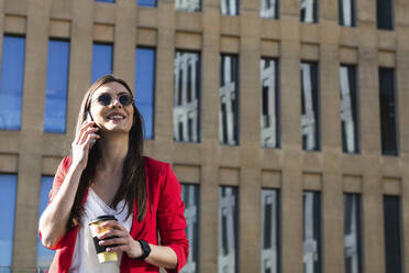 Smiling businesswoman talking on mobile phone while holding coffee cup against building - PNAF00630