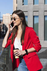 Weibliche Unternehmerin, die mit einem Mobiltelefon spricht und eine Kaffeetasse gegen ein Gebäude hält - PNAF00631