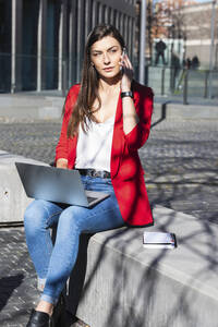 Freiberuflerin mit Laptop, die auf einer Stützmauer sitzend mit ihrem Mobiltelefon spricht - PNAF00633