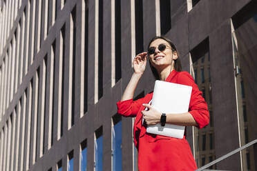 Smiling businesswoman with sunglasses holding laptop while standing against building - PNAF00634