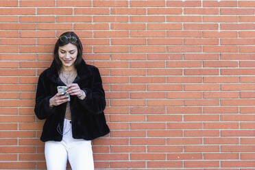Smiling woman with mobile phone listening music while standing against brick wall - PNAF00649