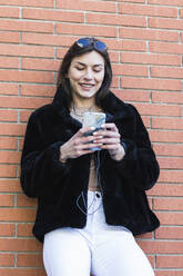 Lächelnde Frau mit Handy hört Musik und lehnt sich an eine Mauer - PNAF00654